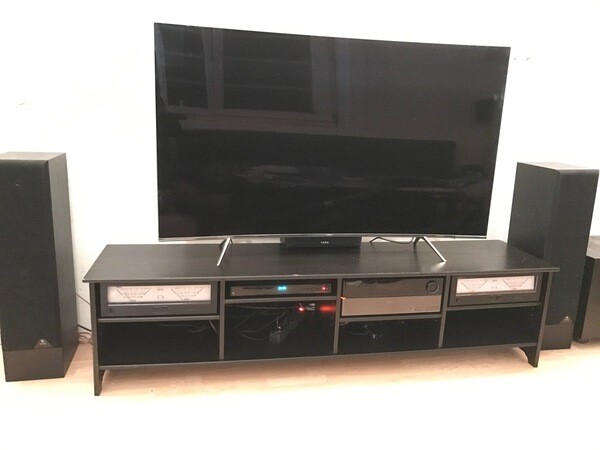 Flachbildfernseher auf schwarzer TV-Konsole, flankiert von zwei Standlautsprechern im Wohnzimmer.