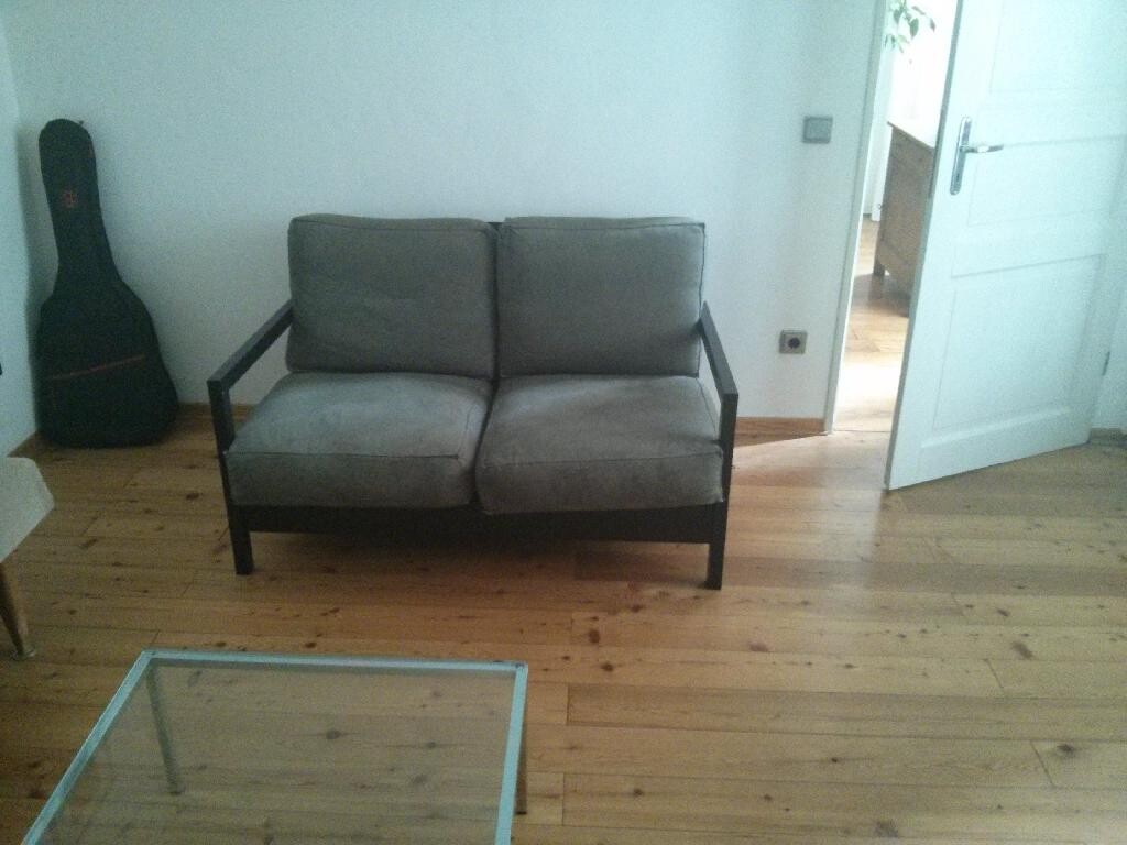 Wohnzimmer mit grauem Sofa auf Holzboden, Glastisch im Vordergrund und Gitarrenkoffer an der Wand.