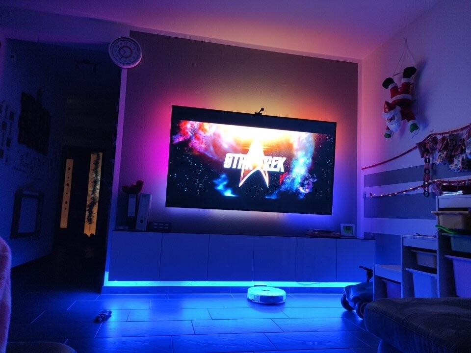 Wohnzimmer mit großem Fernseher, blau-lila LED-Beleuchtung und Roboter-Staubsauger.