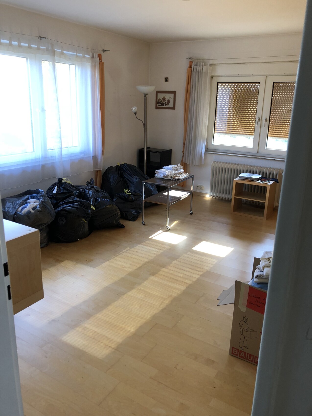 Wohnzimmer mit schwarzen Müllsäcken am Boden, Stehlampe, Tisch, Fenster mit Rollläden.