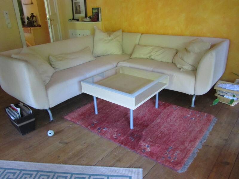 Helles Wohnzimmer mit weißem Ecksofa, rotem Teppich und Couchtisch vor gelber Wand.