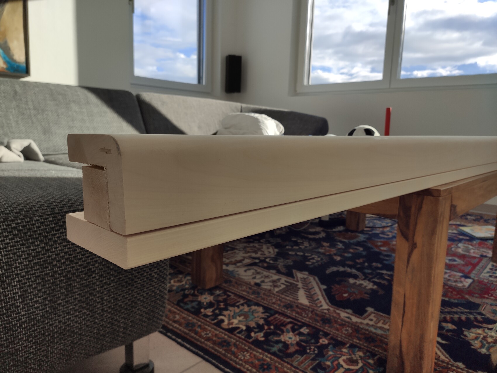 Längliches helles Holzbrett liegt quer über zwei Holzbalken; Sofa und Fenster sind im Hintergrund.