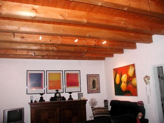 Wohnzimmer mit Holzbalkendecke, dunklem Sofa und bunten Wandbildern.