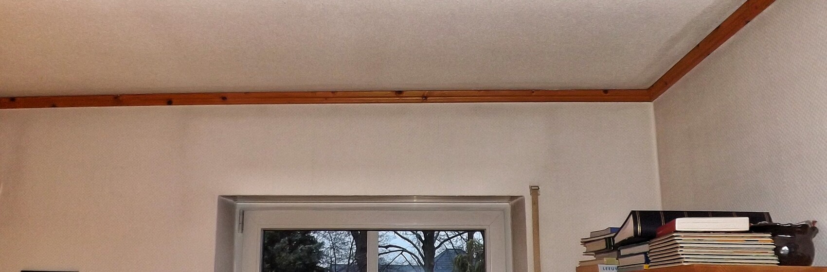 Einrichtung eines Wohnzimmers mit Fenster, Holzbalken an der Decke und Bücherstapel auf einem Regal.