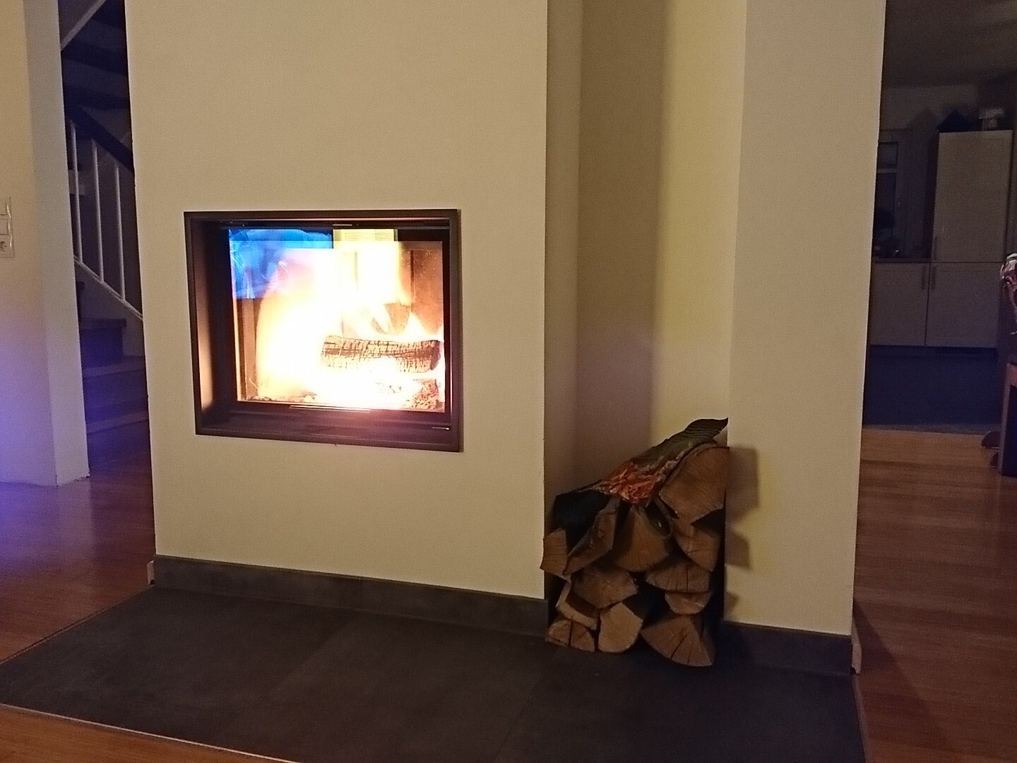Eingebauter Kamin in einer hellen Wand, Flammen lodern; rechts daneben gestapeltes Brennholz.