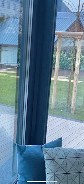 Blick durch eine große Glas-Schiebetür auf Terrasse, Rasen und Garten; Innenkissen am Sofa.