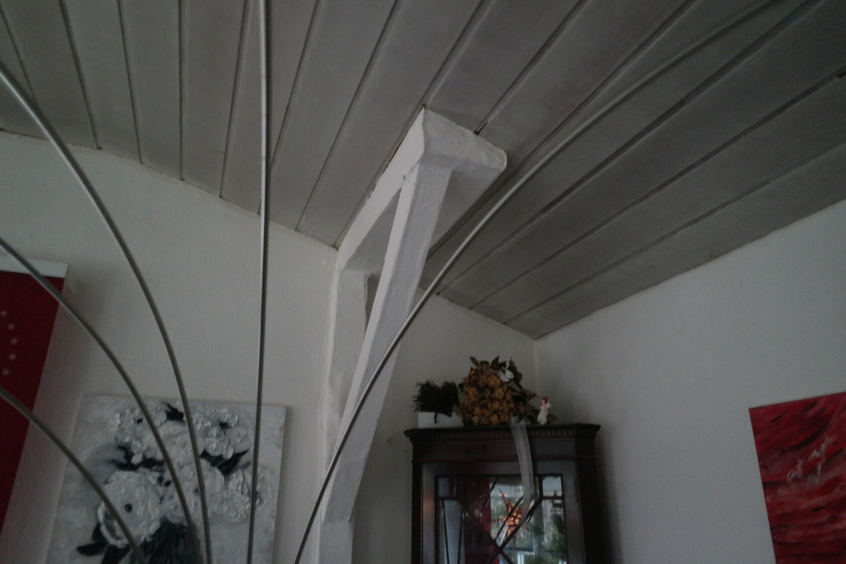 Innenraumecke: weiße diagonale Stütze, graue Deckenpaneele, dunkler Vitrinenschrank mit Dekoration.