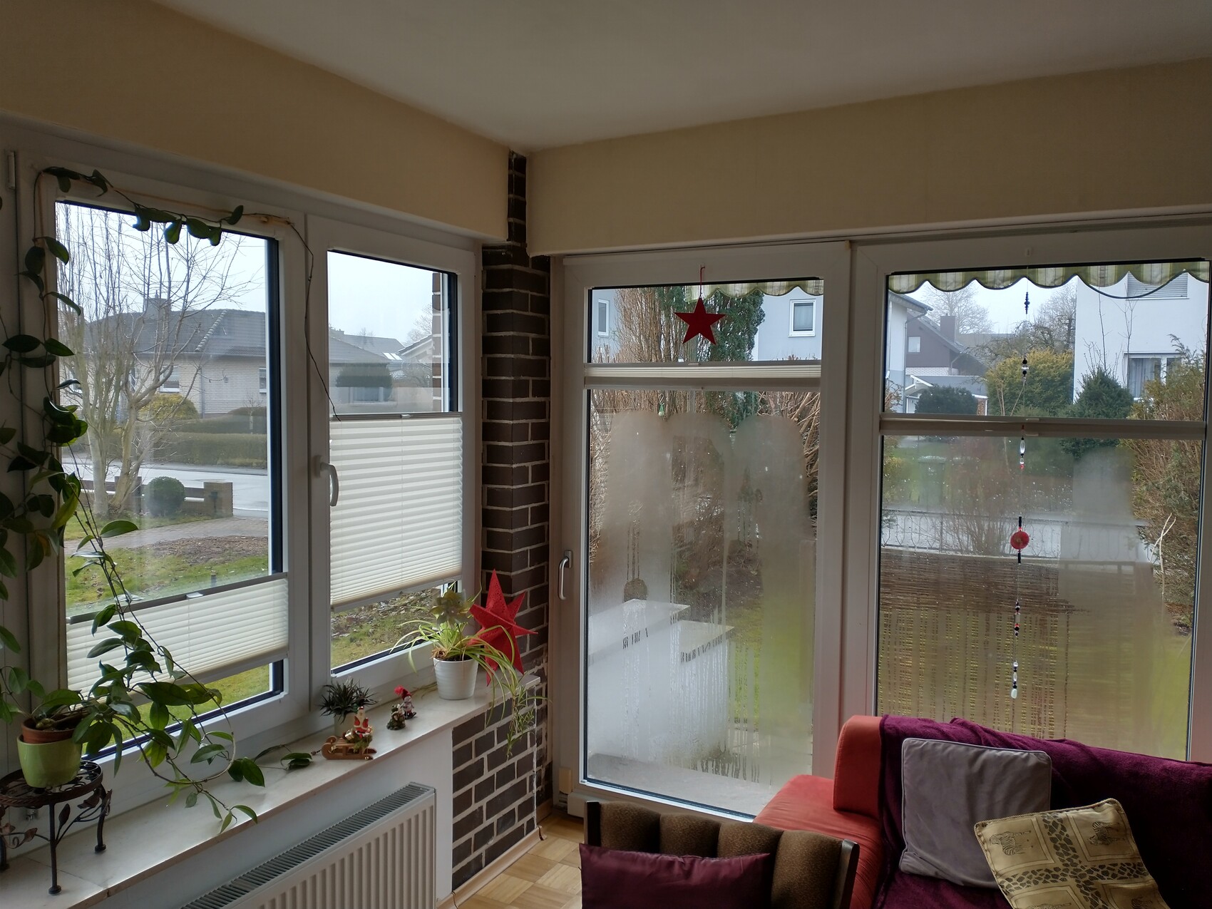 Wohnzimmer mit großen Fenstern, Fensterbankpflanzen und rotem Sternanhänger; Blick nach draußen.