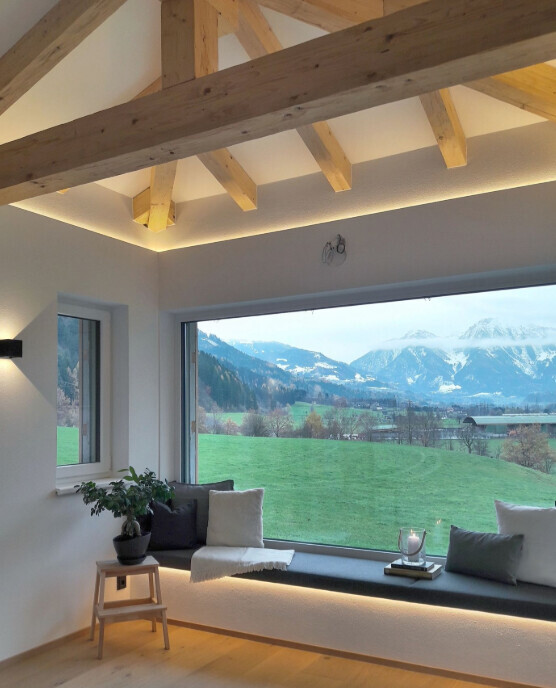 Wohnzimmer mit Fensterfront, Blick auf grüne Wiesen und Berge; Holzdecke, Sitzbank mit Kissen.