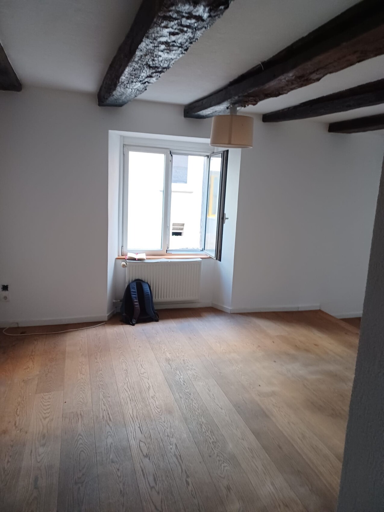 Heller, leerer Raum mit Holzdielen, dunklen Deckenbalken, offenem Fenster und Rucksack.