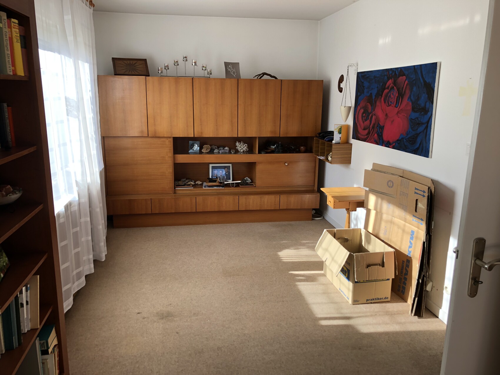 Wohnzimmer mit großem Holzschrank, Kartons rechts und Rosenbild an der Wand.