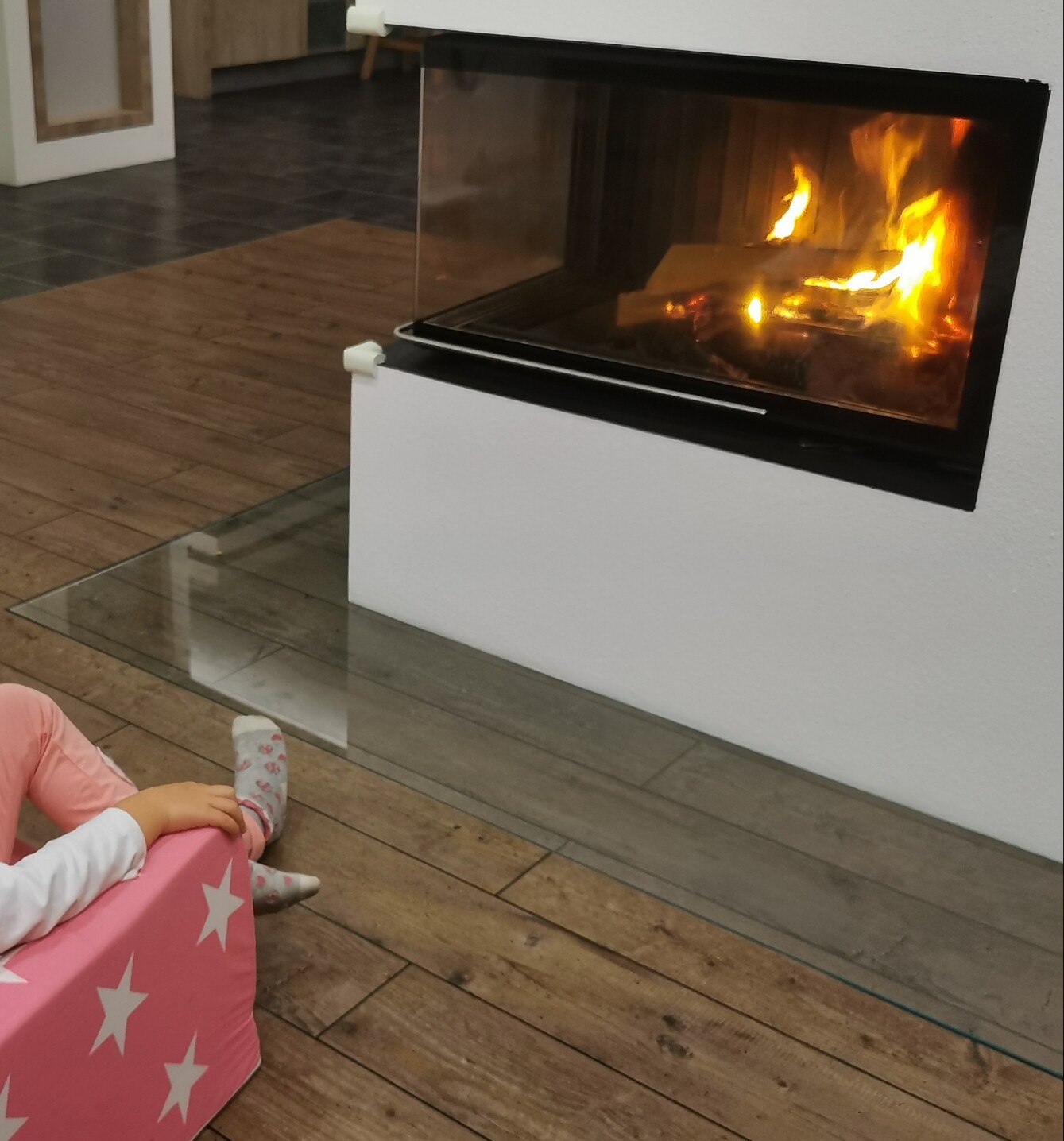 Moderner Kamin in weißer Wand mit Glasfront und brennendem Feuer; Kind auf pinkem Sternen-Sessel.