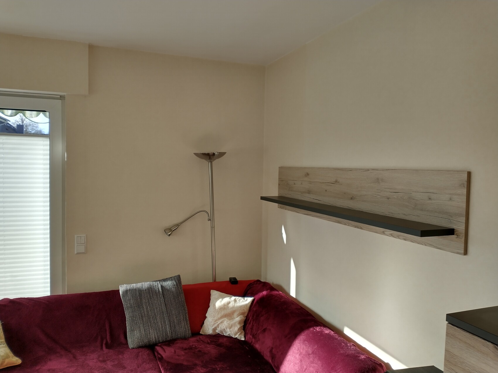 Wohnzimmer-Ecke: mit rotem Sofa, Kissen, Stehleuchte, Wandregal und Fenster mit Blenden.