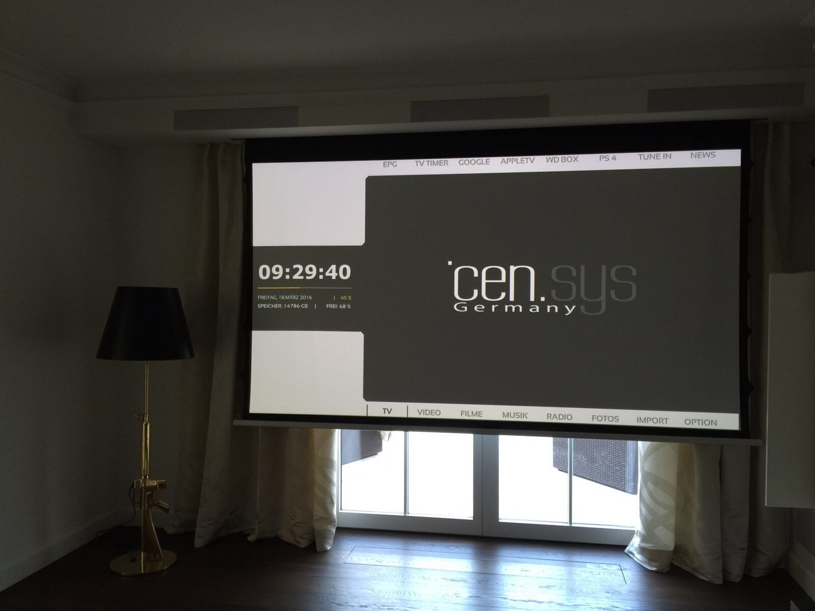 Projektionswand im Wohnzimmer zeigt Mediacenter-UI und Uhrzeit 09:29:40; Lampe links.