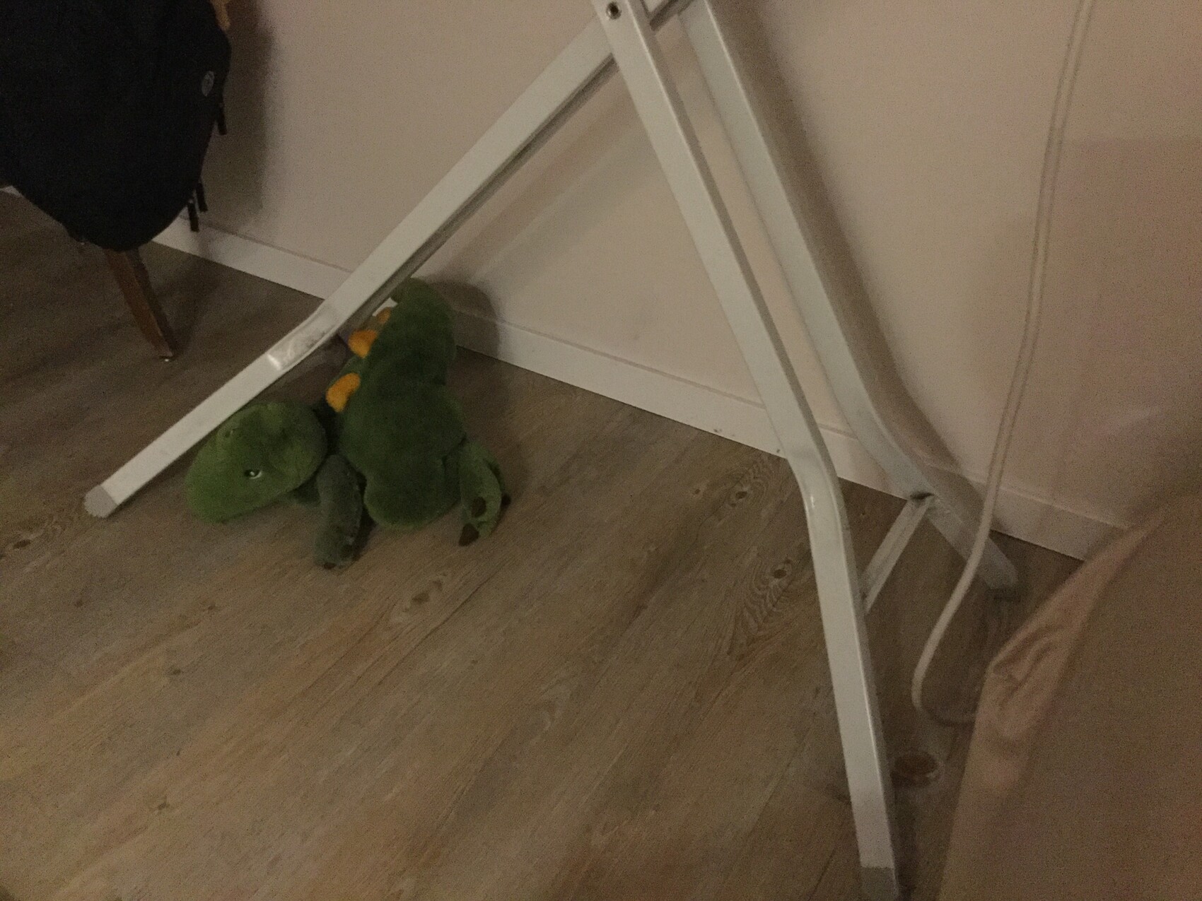 Grüner Plüsch-Dino mit orange Streifen liegt unter einem weißen Klapptisch auf Holzboden.