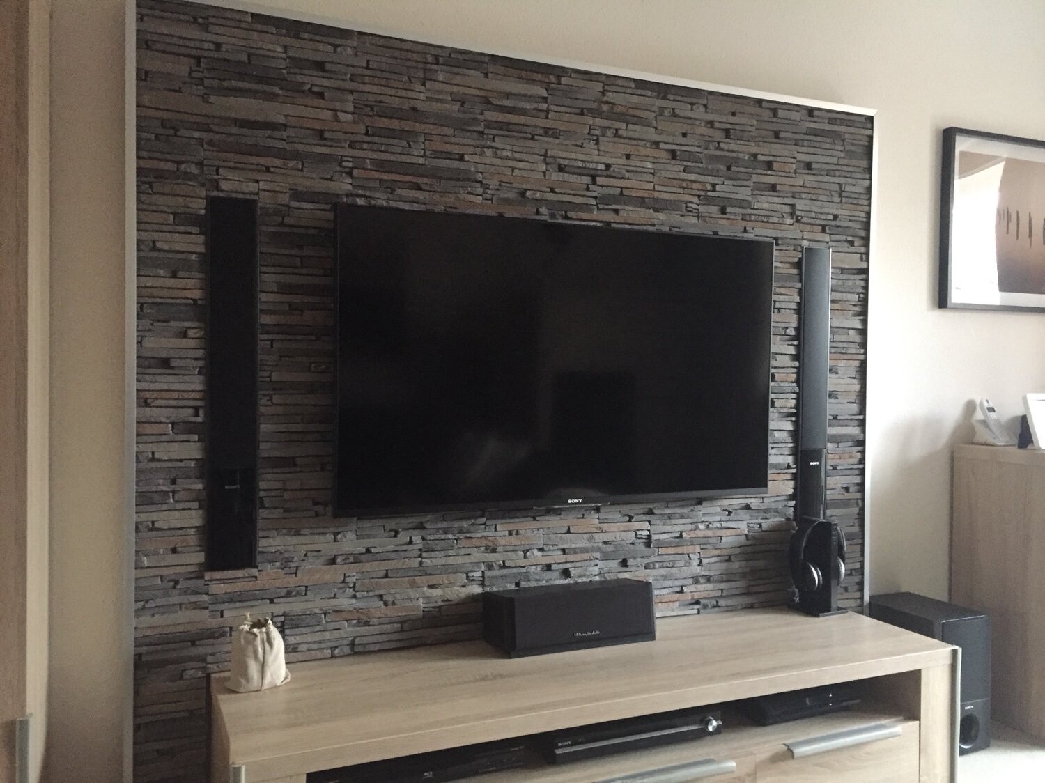 Großer Flachbildfernseher an Steinwand, flankiert von vertikalen Lautsprechern; Soundbar unten.