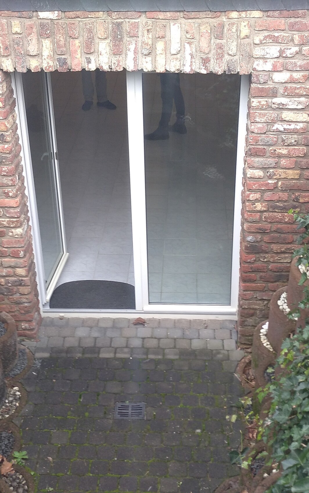 Glas-Schiebetür vor roter Backsteinwand; Fliesenboden, Moos am Pflaster, Beine spiegeln im Glas