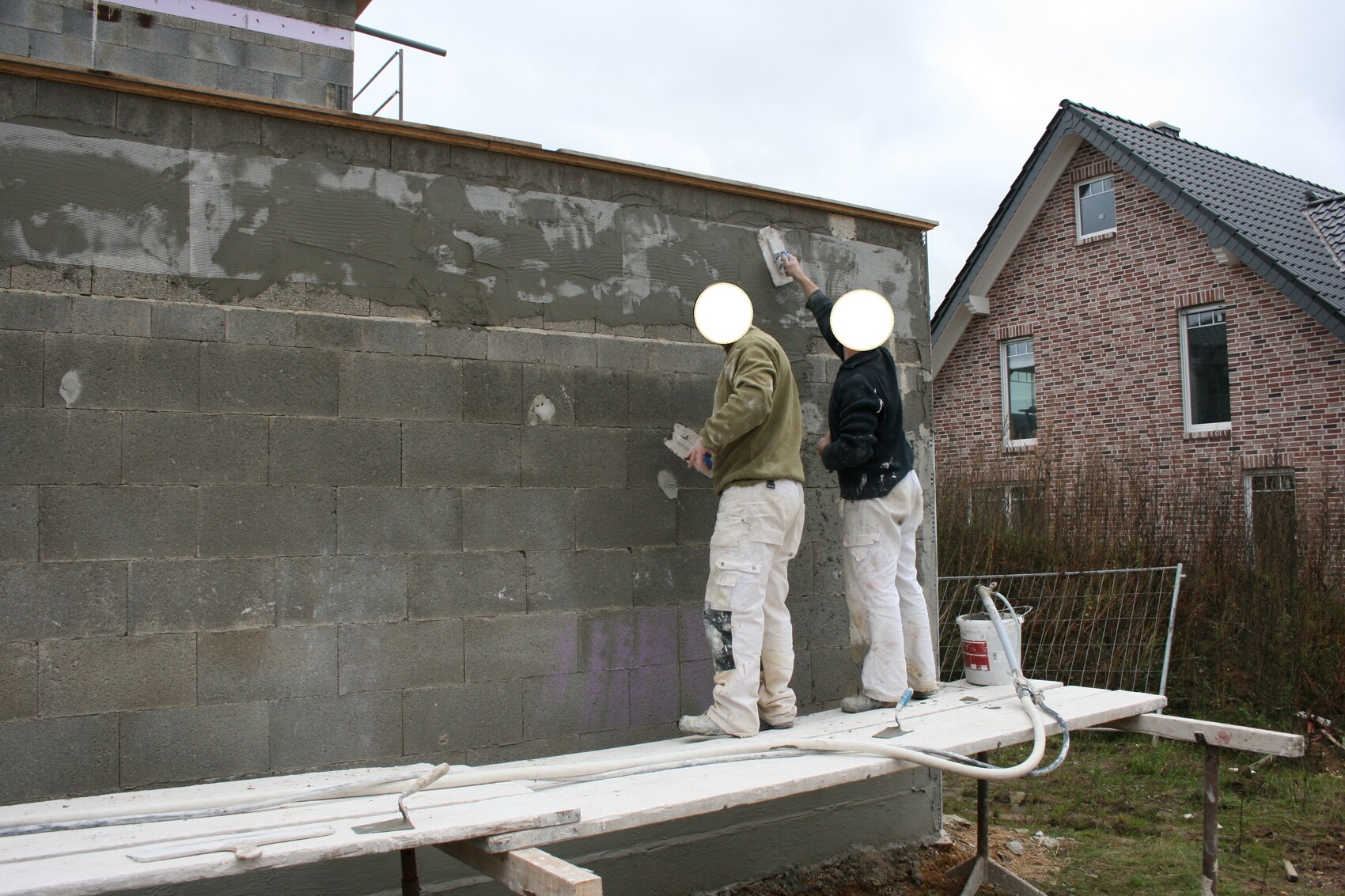 Zwei Bauarbeiter verputzen eine Betonblockwand auf einer Holzbühne; im Hintergrund ein Backsteinhaus.