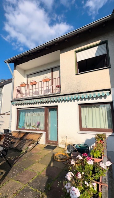 Zweistöckiges Haus mit Balkon, Blumen und Gartenzubehör vor Eingang, blauer Himmel.
