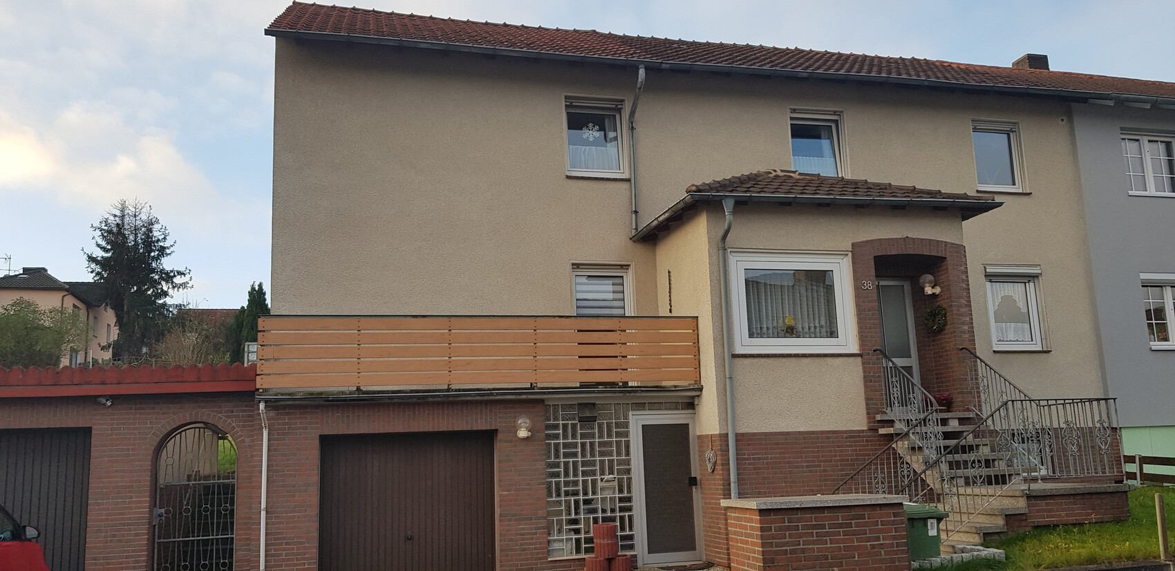 Beige zweistöckiges Haus mit rotem Ziegelsockel, Holzbalustrade links und Treppe rechts.