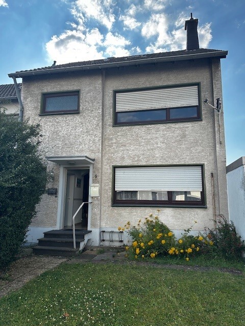 Zweistöckiges Haus mit grauer Putzfassade, zwei Fenster mit Rollläden, Vorgarten mit gelben Blumen.