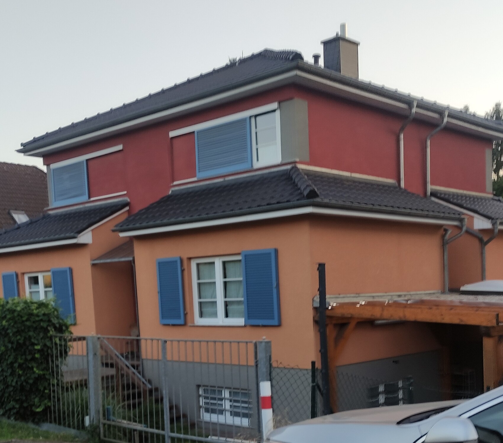 Zweistöckiges Haus mit oranger Front, rotem Obergeschoss, blauen Fensterläden, Gartenzaun und Carport.