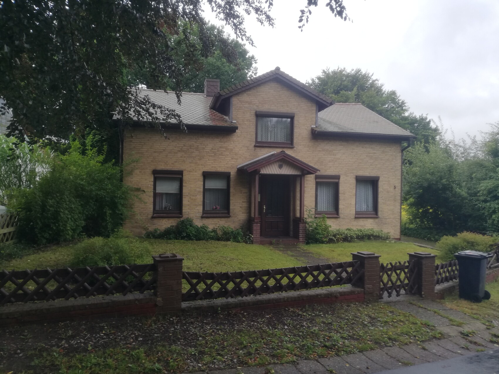 Hellbraunes zweistöckiges Einfamilienhaus mit Vortreppe, braunen Fenstern, Garten und Holzzaun.